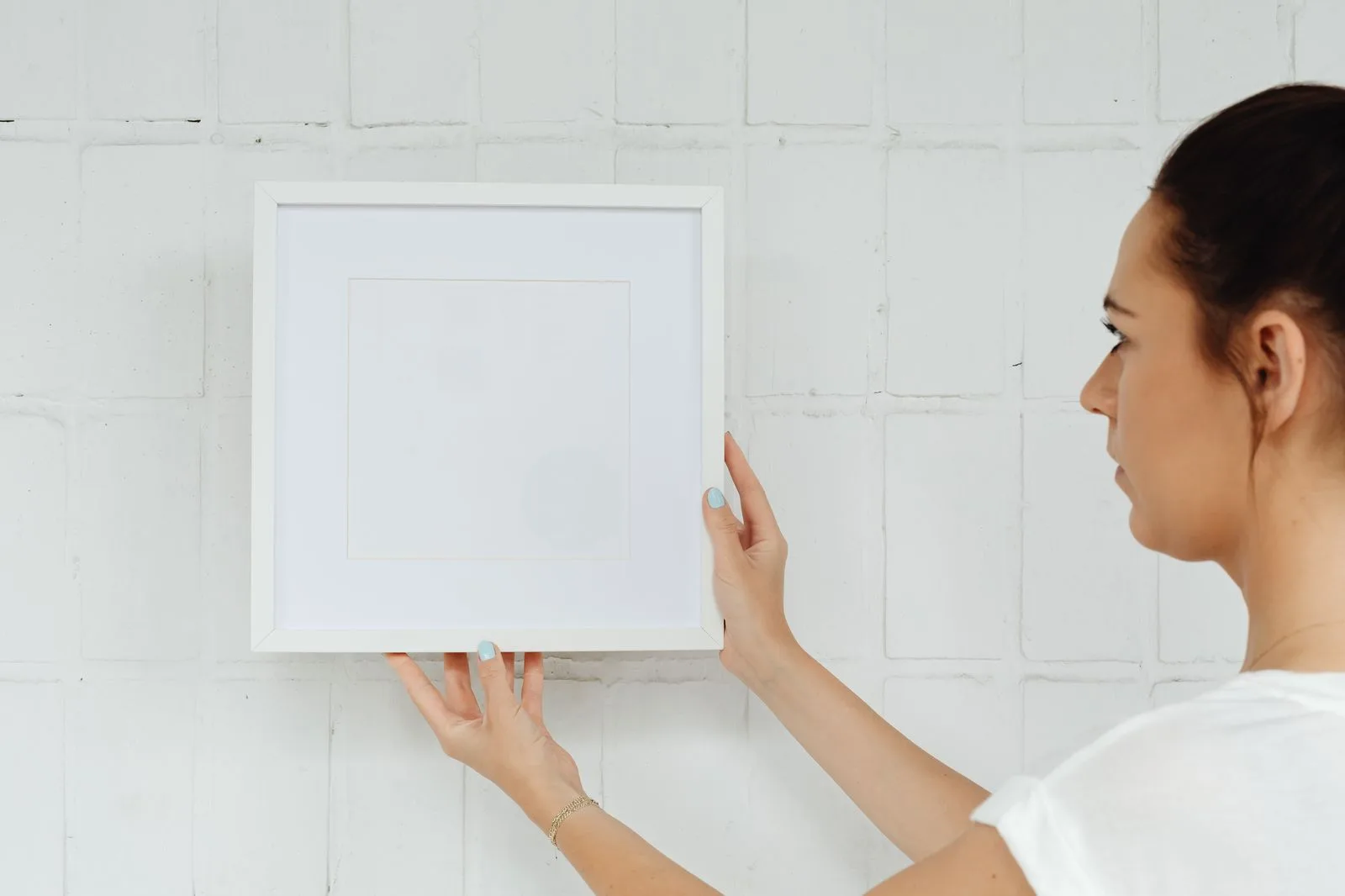 Woman holding a white square frame up against a painted brick wall, ready to hang