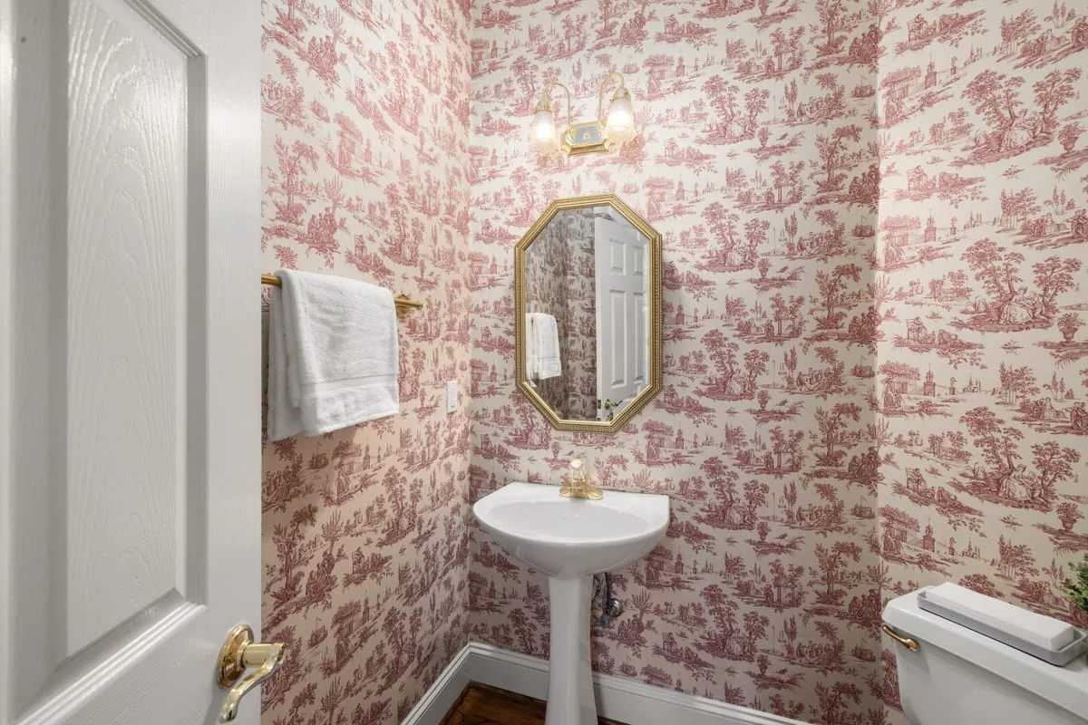 Half-bath powder room with pink toile peel-and-stick wallpaper above a pedestal sink and gold-framed mirror